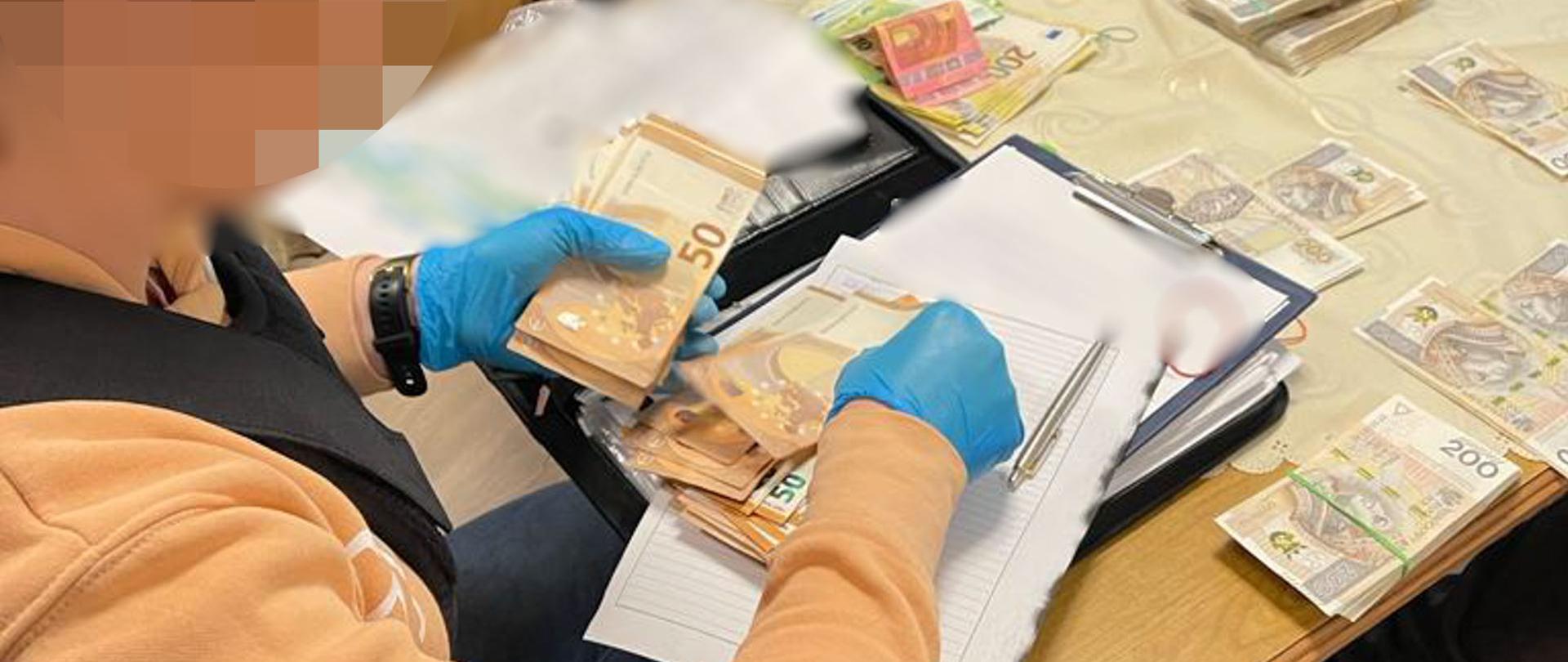 Funkcjonariusz przelicza zatrzymaną gotówkę. Na stole pliki banknotów w różnej walucie