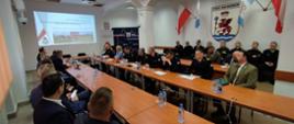 Widok na sale w której odbywa się prezentacja pracy KP PSP Białogard. Na ścianie prezentacja, przy stołach strażacy i goście.