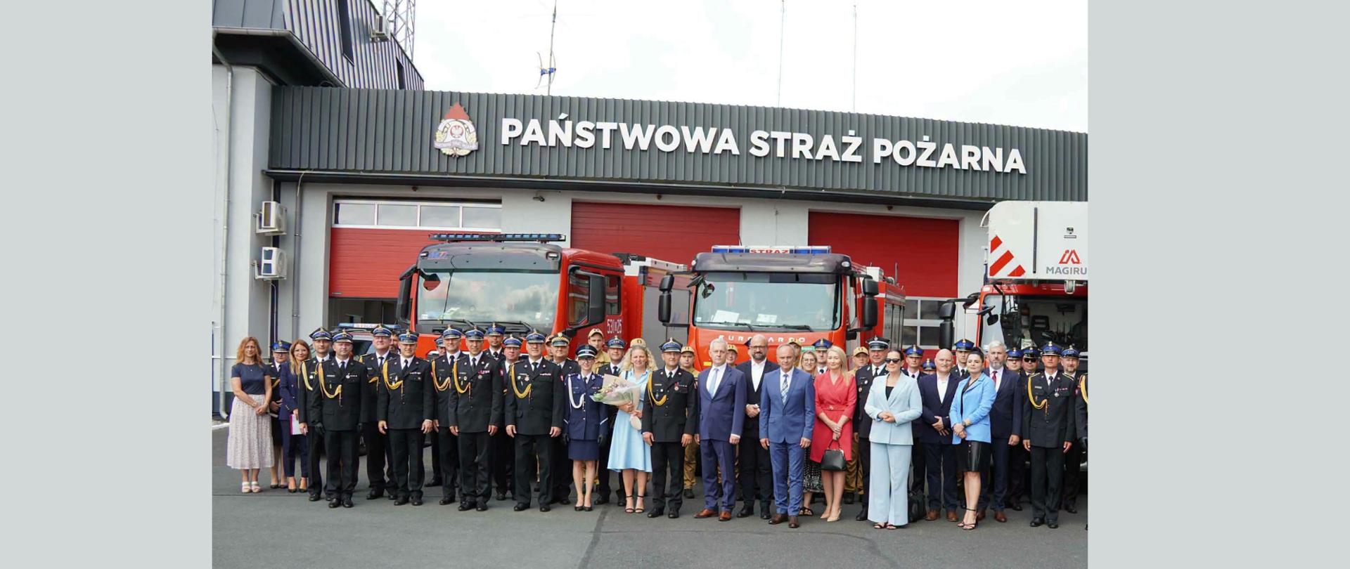 Zdjęcie przedstawia oficjalną, grupową fotografię wykonaną na zewnątrz, przed budynkiem Państwowej Straży Pożarnej. W tle widoczne są duże, czerwone wozy strażackie. Na pierwszym planie stoi grupa około 30-40 osób ustawionych w dwóch rzędach. Są to głównie strażacy w ciemnogranatowych mundurach wyjściowych oraz zaproszeni goście – cywile w eleganckich strojach wizytowych (garniturach i sukienkach). Atmosfera zdjęcia jest oficjalna i uroczysta, prawdopodobnie dokumentuje ono ważne wydarzenie, święto lub przekazanie sprzętu.