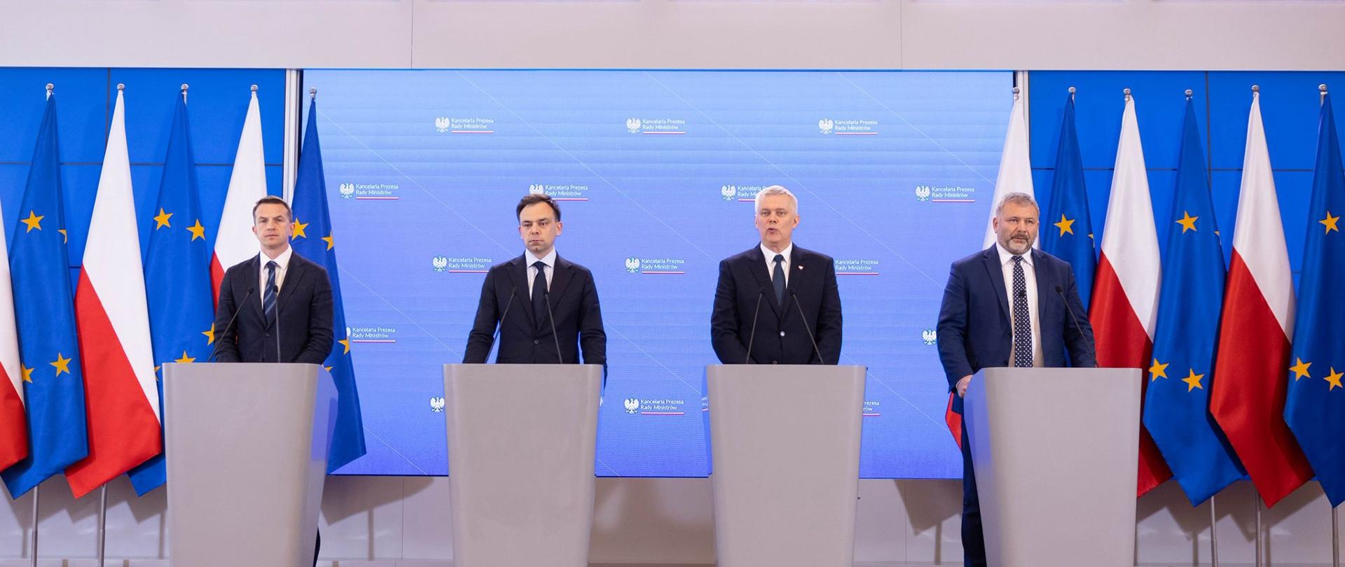 Na pierwszym planie zdjęcia są widoczni, od lewej: Rzecznik rządu Adam Szłapka, Minister Finansów i Gospodarki Andrzej Domański, Minister, Koordynator Służb Specjalnych Tomasz Siemoniak oraz Minister Sprawiedliwości Waldemar Zurek. Stoją za szarymi mównicami. Za ministrami widać niebieski ekran z logo KPRM, po bokach ekranu stoją flagi Polski i Unii Europejskiej.