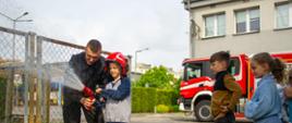 Dziecko w kasku strażackim leje wodę z prądownicy wodnej, pomagając mu strażak podczas pokazu na dniach otwartych strażnicy