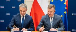 Za stolikiem siedzi minister Wieczorek i mężczyzna w szarym garniturze, obaj trzymają długopisy i podpisują leżące przed nimi dokumenty, za nimi pod ścianą flagi Polski i UE.