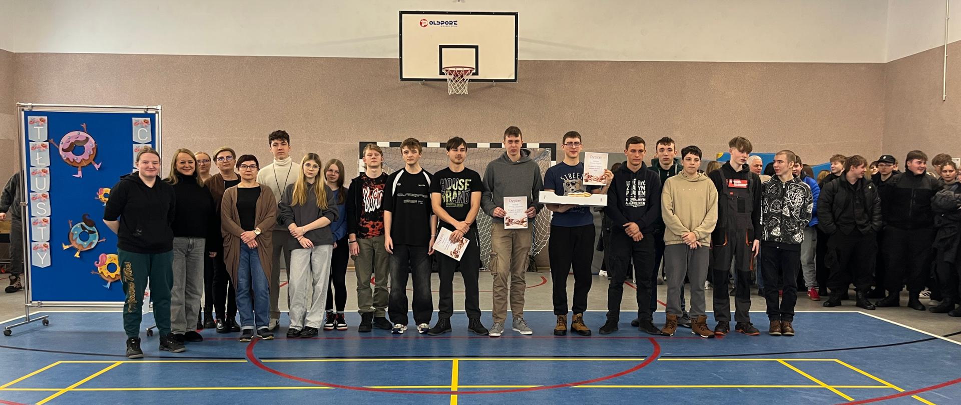 Duża grupa uczniów i nauczycieli stoi na środku hali sportowej z dyplomami oraz pudełkiem pączków, świętując tłusty czwartek. Po lewej stronie widoczna jest niebieska tablica z dekoracjami w kształcie pączków oraz napisem „Tłusty czwartek”.