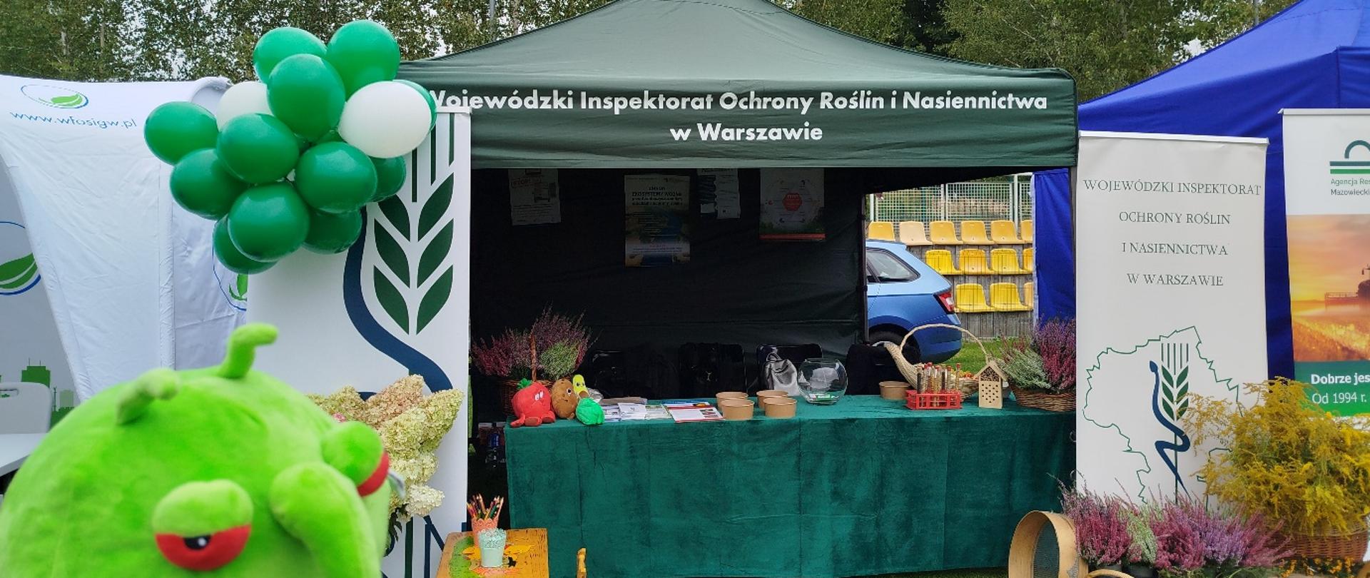 Stoisko plenerowe WIORiN w Warszawie z zielonym namiotem, banerami i dekoracjami z kwiatów, balonów i koszy z owocami; na pierwszym planie widoczna maskotka w kształcie zielonego owada.