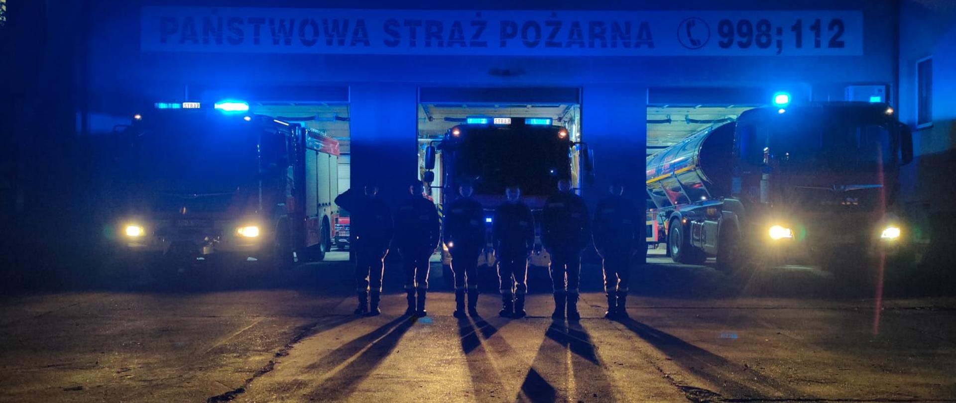 Przed strażackim wozem bojowym stoi kilku strażaków w mundurach jeden z nich salutuje.