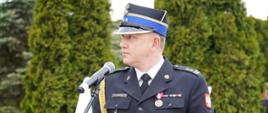 Komendant Powiatowy Państwowej Straży Pożarnej w Rypinie w galowym mundurze wygłasza przemówienie przy mównicy podczas obchodów Dnia Strażaka. Na mundurze widoczne są odznaczenia i medale, a w tle znajdują się biało-czerwone flagi.