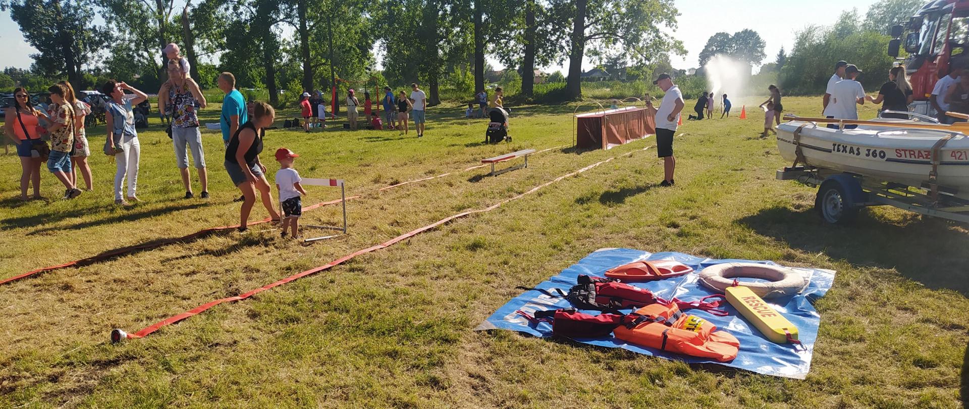W pogodny dzień dzieci z rodzicami uczestniczą w akcji bezpieczne wakacje. W tle tor edukacyjny przygotowany przez strażaków.