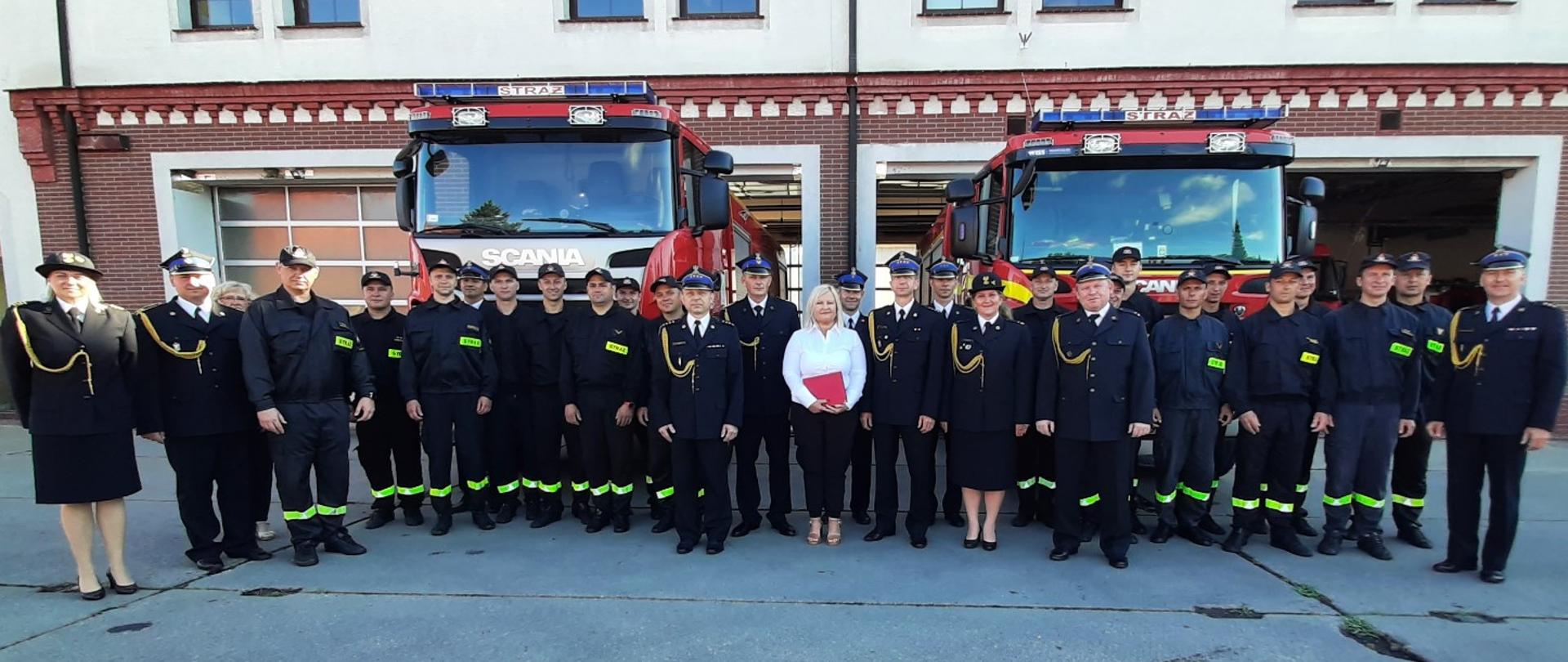 Zdjęcie przedstawia strażaków i osobę cywilną stojące przed samochodami pożarniczymi, w tle budynek strażnicy