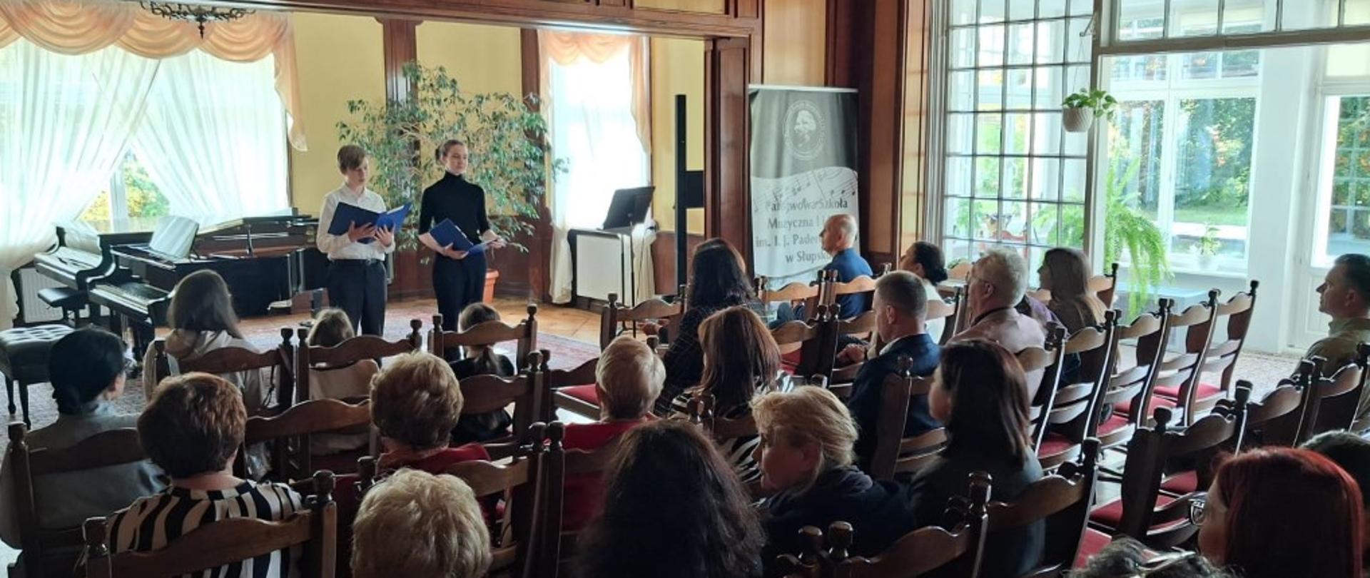 Zdjęcie kolorowe wnętrze sali koncertowej w dworku, w której odbywa się spotkanie i koncert. Na ścianie za sceną polski herb – biały orzeł w czerwonym polu. Dwoje uczniów chłopców na ) scenie, jeden z nich stoi w centrum i trzyma niebieski folder, drugi stoi po jego lewej stronie i trzyma otwarty segregator. W głębi po lewej stronie fortepian. Poniżej na wprost rzędy krzeseł, na których siedzi publiczność tyłem do obiektywu. Po prawej stronie duże okno z widokiem na taras z zimowym ogrodem.
