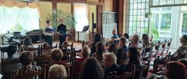 Zdjęcie kolorowe wnętrze sali koncertowej w dworku, w której odbywa się spotkanie i koncert. Na ścianie za sceną polski herb – biały orzeł w czerwonym polu. Dwoje uczniów chłopców na ) scenie, jeden z nich stoi w centrum i trzyma niebieski folder, drugi stoi po jego lewej stronie i trzyma otwarty segregator. W głębi po lewej stronie fortepian. Poniżej na wprost rzędy krzeseł, na których siedzi publiczność tyłem do obiektywu. Po prawej stronie duże okno z widokiem na taras z zimowym ogrodem.