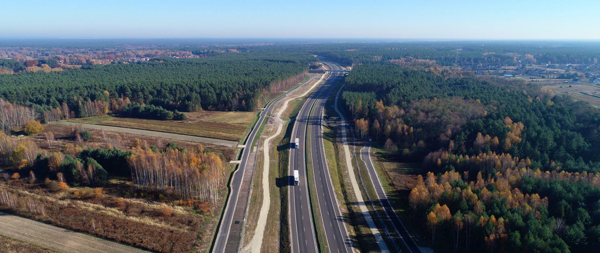 Widok na odcinek drogi ekspresowej S19. Dwie jezdnie po dwa pasy ruchu oddzielone pasem zieleni. Droga przebiega przez tereny leśne. Drzewa w kolorach jesieni.