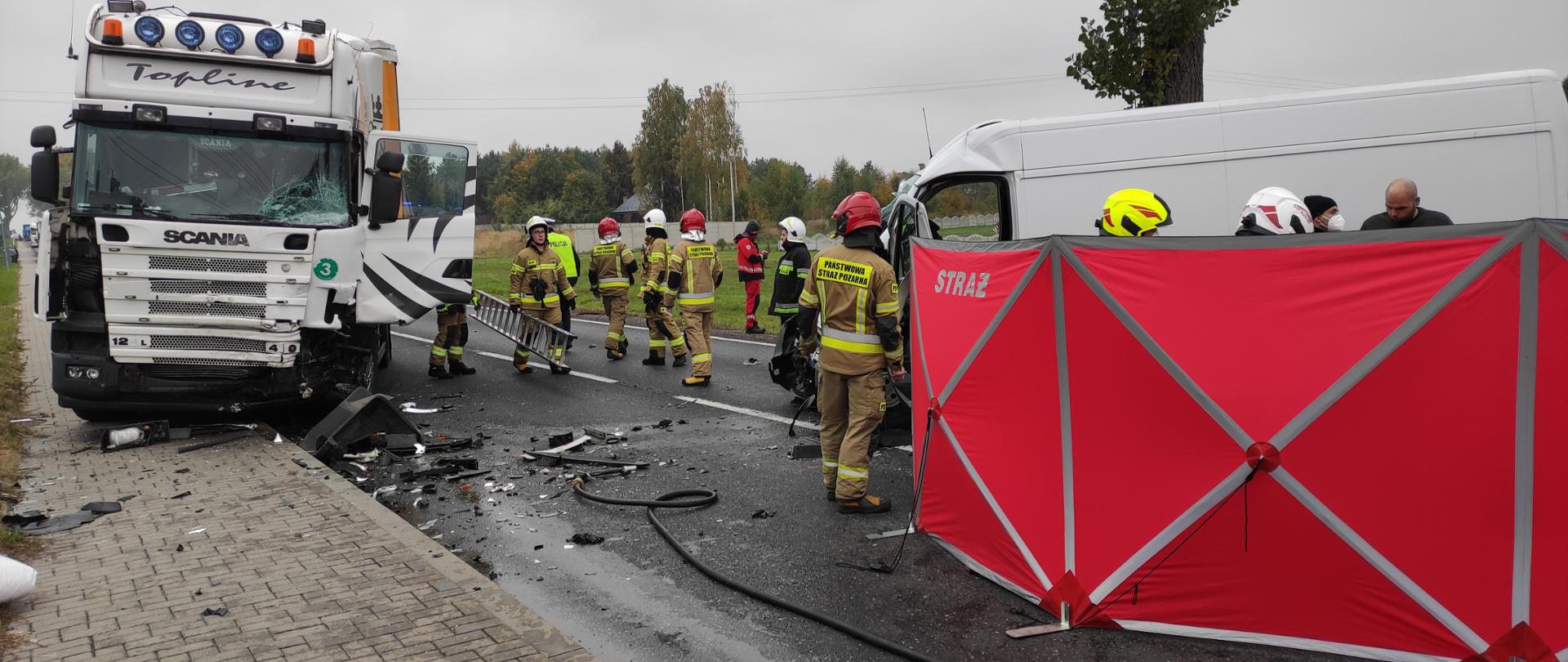 Na zdjęciu samochód ciężarowy koloru białego z uszkodzoną karoserią, stoi częściowo na chodniku a częściowo na drodze. Obok strażacy w hełmach oraz ubraniach specjalnych. Po prawej stronie na drodze rozstawiony parawan koloru czerwonego z tył widać białego busa.