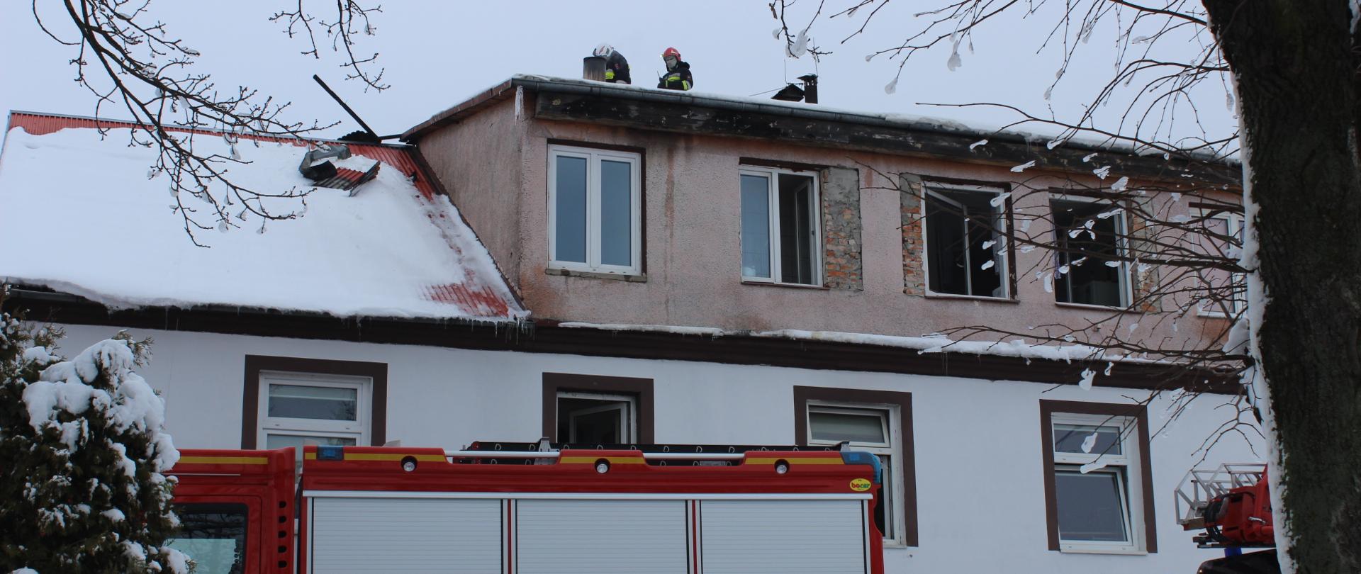 Front budynku handlowo-usługowego. Na dachu pracujący strażacy. Przed budynkiem stoi samochód ratowniczo-gaśniczy.