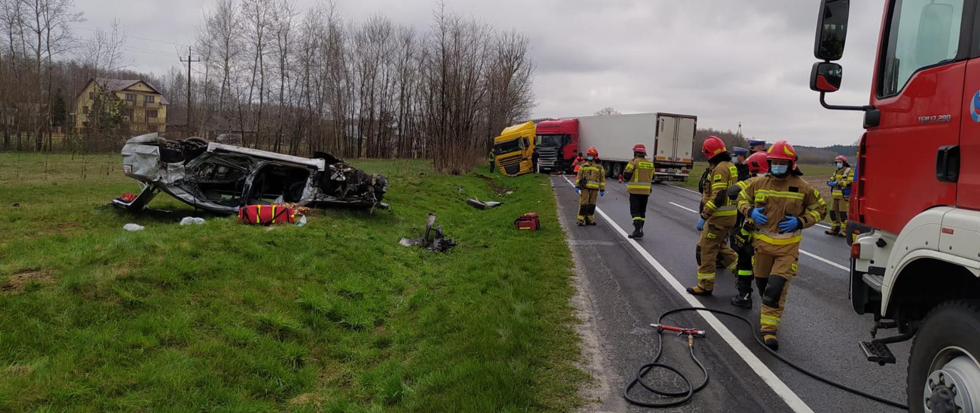 Na zdjęciu widać pracujących strażaków, część wozu strażackiego oraz sprzęt. W rowie znajduje się pognieciony samochód osobowy a w oddali widać dwie ciężarówki, które uległy zderzeniu.