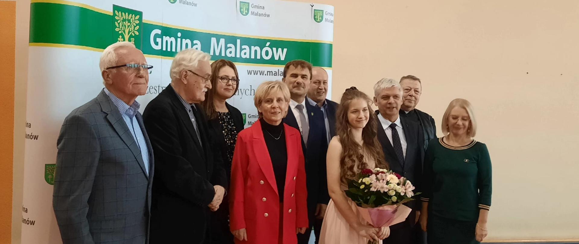 Zdjęcie przedstawia uczestników i prelegentów uroczystości w Malanowie, którzy stoją przed banerem Gmina Malanów ubrani na galowo. Pianistka trzyma bukiet kwiatów.