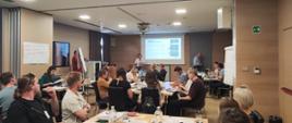 Sala konferencyjna podczas warsztatów lub szkolenia. Kilkadziesiąt osób siedzi przy stołach ustawionych w grupach i uczestniczy w spotkaniu. Na środku sali prelegentka prowadzi prezentację wyświetlaną na ekranie. Uczestnicy korzystają z laptopów, notatek i materiałów szkoleniowych. W pomieszczeniu znajdują się tablice typu flipchart oraz materiały do pracy warsztatowej.