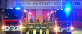 Widoczni strażacy na tle pojazdów gaśniczych i garaży, oddają honor