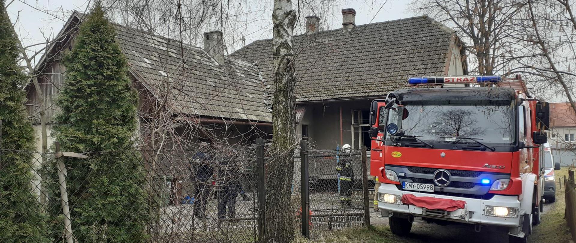 Zdjęcie wykonane na zewnątrz z za ogrodzenia obejmujące budynek mieszkalny. W środkowej części widoczne jest drzewo oraz postacie stojące obok domu. Z prawej strony stoi z włączonymi światłami czerwony samochód strażacki, za nim widać małą część karetki pogotowia ratunkowego.
