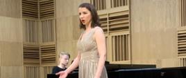 Przy fortpeianie, na scenie stoi młoda dziewczyna w pięknej wieczorowej sukni. Śpiewa do akompaniamentu fortepianu.