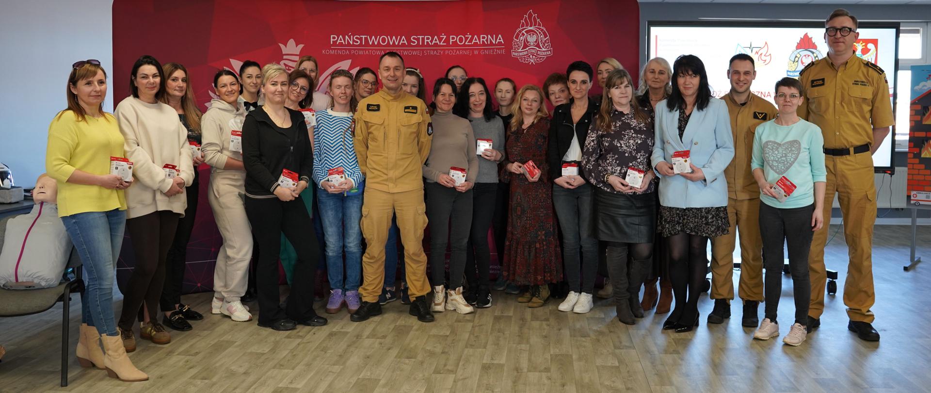 Grupa kilkunastu osób stoi w pomieszczeniu szkoleniowym na tle czerwonej ścianki z napisem „Państwowa Straż Pożarna”. Wśród nich znajdują się strażacy w żółtych mundurach oraz uczestnicy szkolenia – głównie kobiety w codziennych ubraniach. Wszyscy pozują do wspólnego zdjęcia i trzymają w rękach małe czerwono-białe pudełka, prawdopodobnie z czujnikami bezpieczeństwa. Po lewej stronie widać fantom do ćwiczeń pierwszej pomocy, a po prawej ekran prezentacyjny z grafikami związanymi z bezpieczeństwem i pożarnictwem. Wnętrze jest jasne, z dużą salą, panelowym sufitem i drewnianą podłogą.