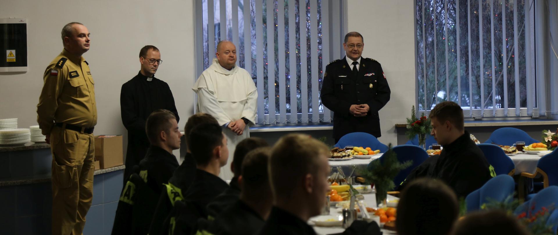 W głębi od lewej stojący oficer CS PSP, ksiądz, paulin oraz Zastępca Komendanta Centralnej Szkoły PSP przemawiający podczas wigilijnego spotkania, przed nimi siedzący za stolami kadeci CS PSP