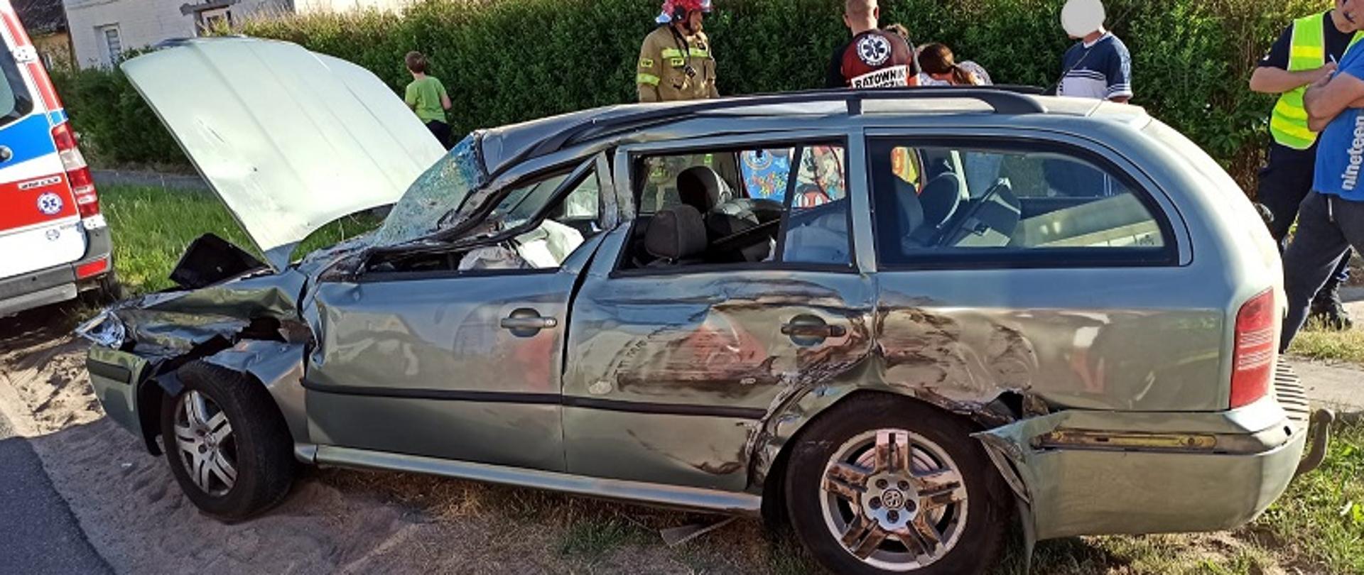 Wypadek w miejscowości Słonino w powiecie białogardzkim. Zdjęcie przedstawia stojący na poboczu widoczny bokiem od strony kierowcy szary samochód marki Skoda Octavia. Karoseria pojazdu jest uszkodzona, szyba przednia pobita. Za samochodem strażacy, ratownicy medyczni, policjanci i osoby uczestniczące w wypadku