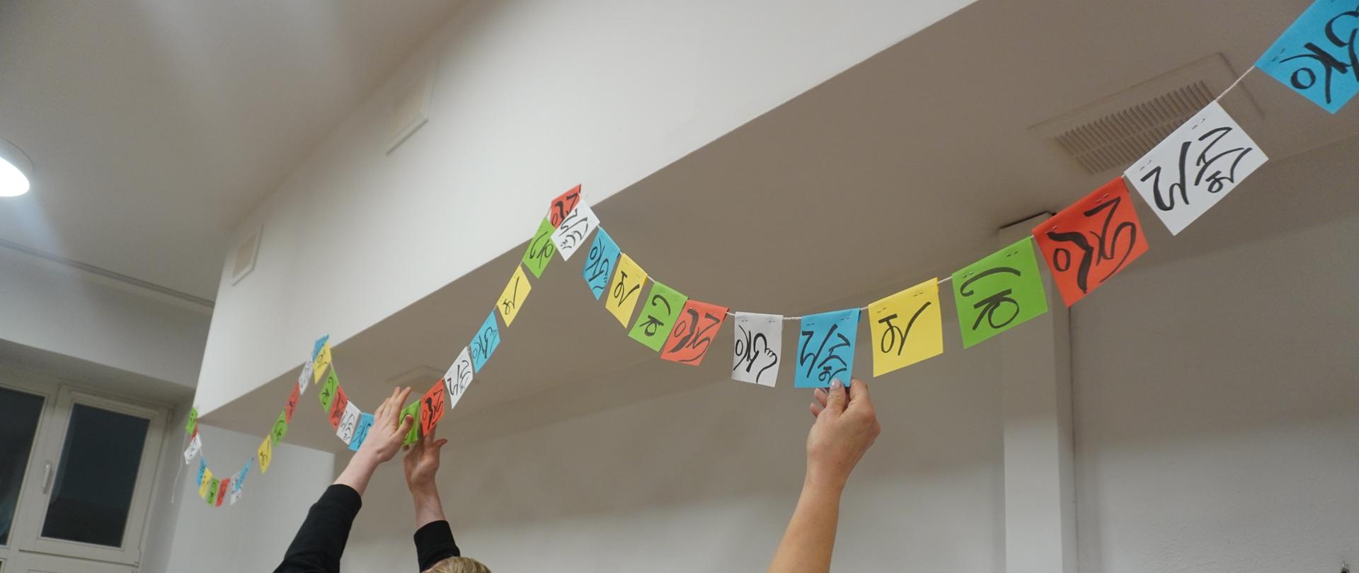 Pani Katarzyna i wychowanek rozwieszają dekoracje w stołówce z barwami i alfabetem nepalskim