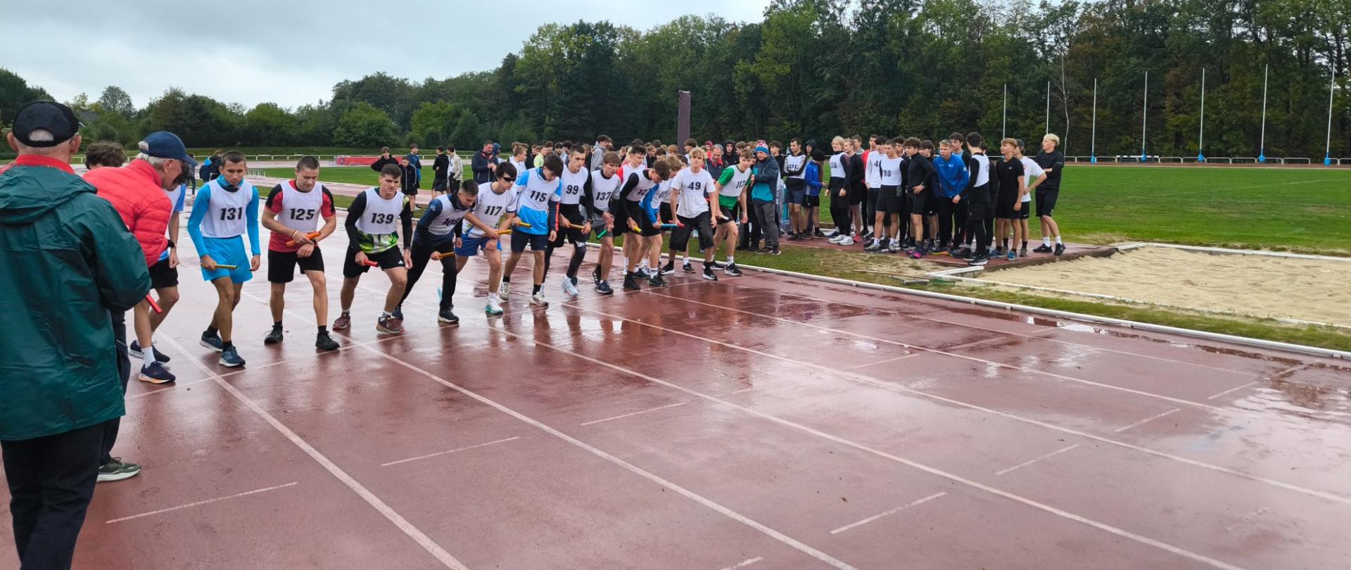 Zawodnicy z numerami 131, 125, 139, 117 i 99 stoją na mokrej bieżni na linii startu biegu. Część jest w pozycji startowej, inni czekają za nimi. W tle widoczne są trawa, drzewa i piaskownica do skoku w dal.