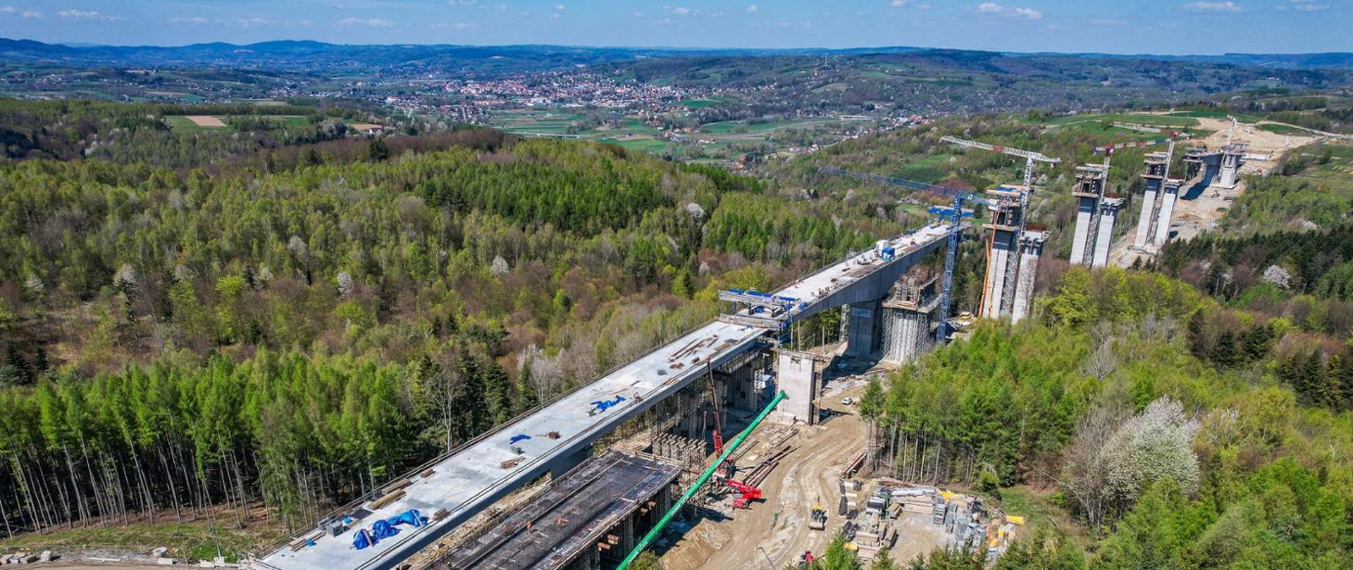 Zdjęcie wykonane z lotu ptaka przedstawia rozległy plac budowy drogi ekspresowej S19 w górzystym, leśnym terenie. Przez środek kadru biegnie bardzo długa estakada na wysokich, masywnych betonowych filarach, częściowo już z wykonaną płytą jezdni. Pod estakadą widoczne są rusztowania, konstrukcje tymczasowe oraz intensywnie prowadzone prace budowlane. Po prawej stronie znajdują się wysokie podpory kolejnych przęseł w trakcie realizacji, a obok nich dźwigi i maszyny budowlane. Na dole widać drogi technologiczne, zaplecze budowy oraz składowane materiały. Całość otoczona jest gęstymi lasami i pagórkami, a w oddali rozciąga się panorama miejscowości i dolin, sięgająca aż po horyzont.
