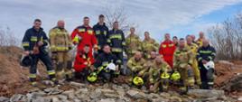 Uczestnicy szkolenia KSRG z zakresu działań poszukiwawczo-ratowniczych pozują do wspólnego zdjęcia po zakończeniu ćwiczeń terenowych. Druhowie OSP z powiatu opolskiego, ubrani w mundury strażackie i ratownicze, stoją na tle gruzowiska, symbolizując gotowość do niesienia pomocy w sytuacjach kryzysowych.