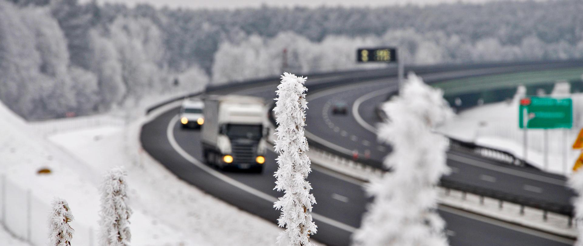 Zdjęcie przedstawia zakręt na drodze ekspresowej S3. Nawierzchnia drogi jest odśnieżona i czarna. Po drodze poruszają się różne pojazdy. Cały teren poza drogą pokryty jest warstwą śniegu. Na pierwszym planie roślina pokryta gęsto szronem.