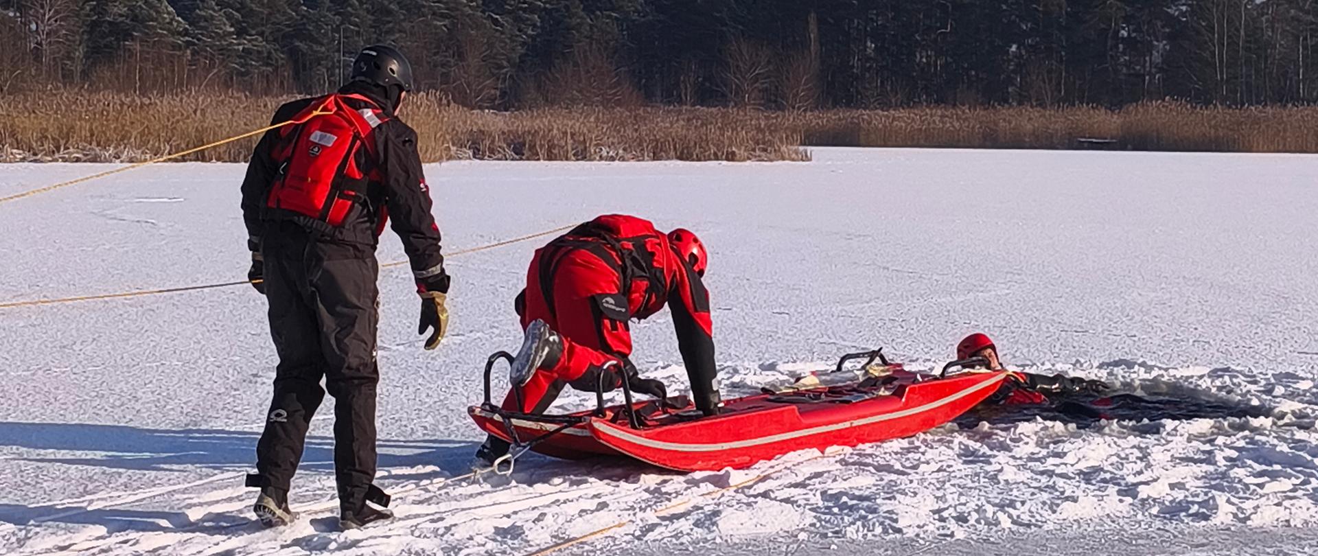 Strażacy w kombinezonach do ratownictwa wodnego przy użyciu sań lodowych podejmują osobę poszkodowaną (pozoranta) z przerębla w lodzie.