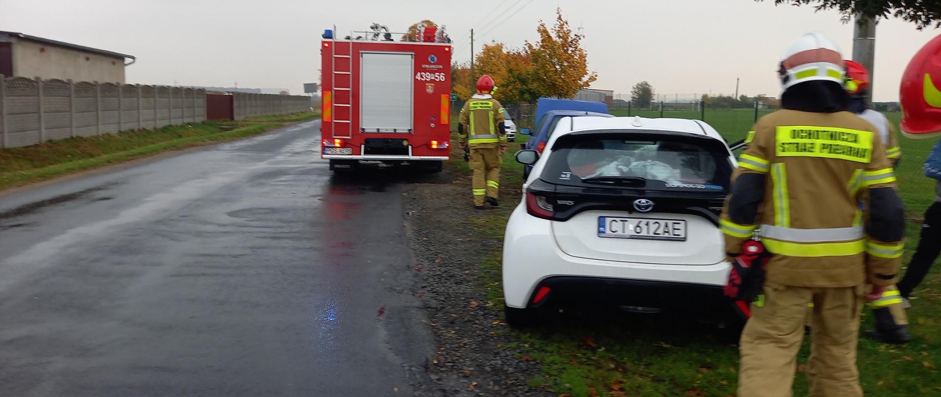 Uszkodzone pojazdy na poboczu drogi. W tle pojazd pożarniczy.