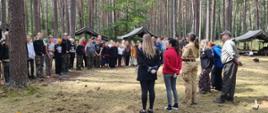 Na zdjęciu wykonanym w obozie harcerskim w lesie znajdują się przedstawiciele PSP oraz służ i instytucji współpracujących prowadzących działania profilaktyczne. Odbiorcami treści związanych z bezpieczeństwem są młodzi harcerze, uczestnicy obozu.