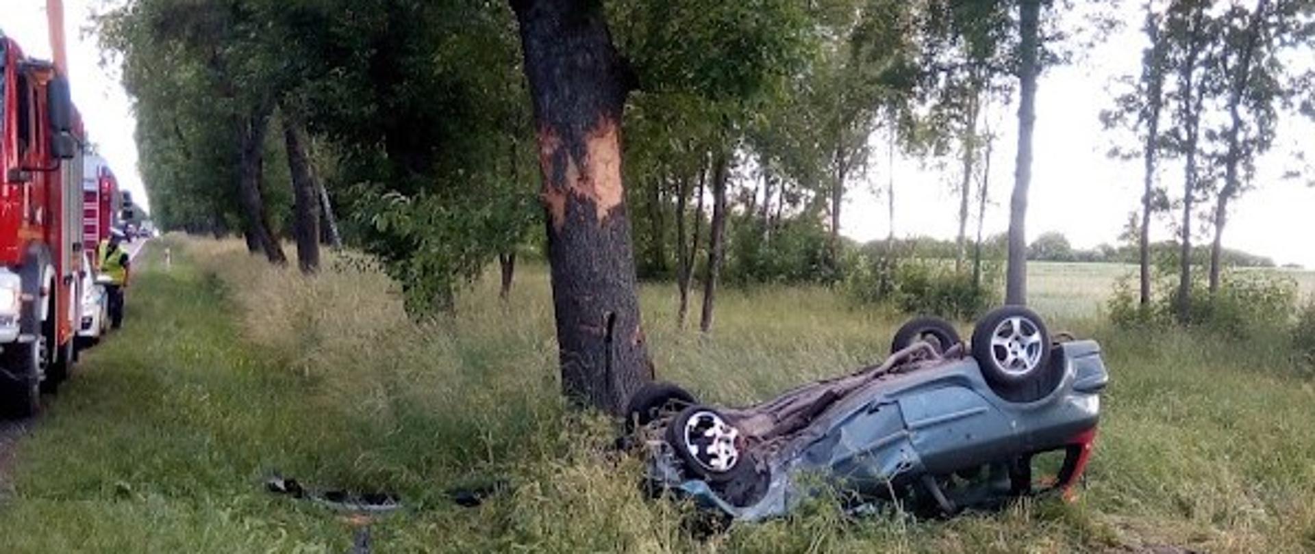 Uszkodzony samochód osobowy leżący na dachu w przydrożnym rowie. Na drodze widoczne samochody ratowniczo-gaśnicze.