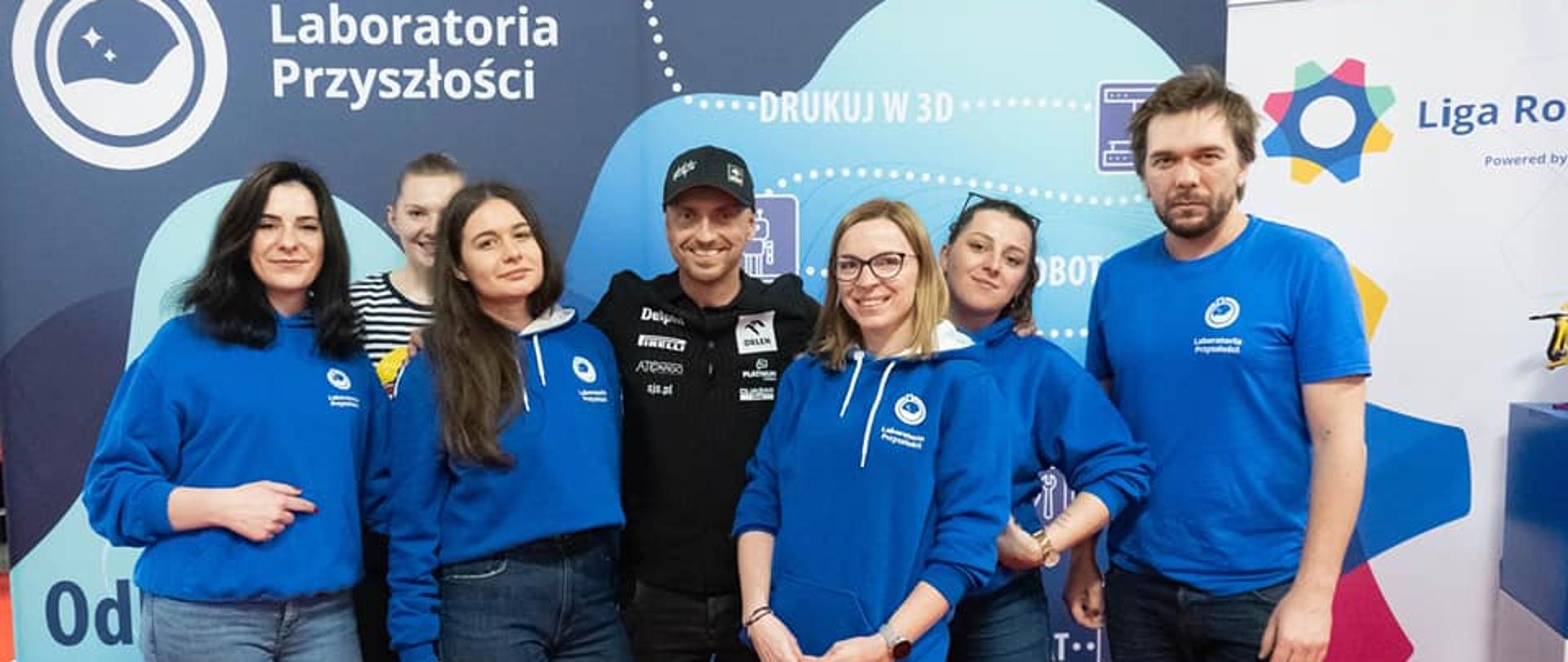 Przed niebieską ścianką z napisem Laboratoria przyszłości stoi grupa uśmiechniętych młodych osób w niebieskich bluzach.