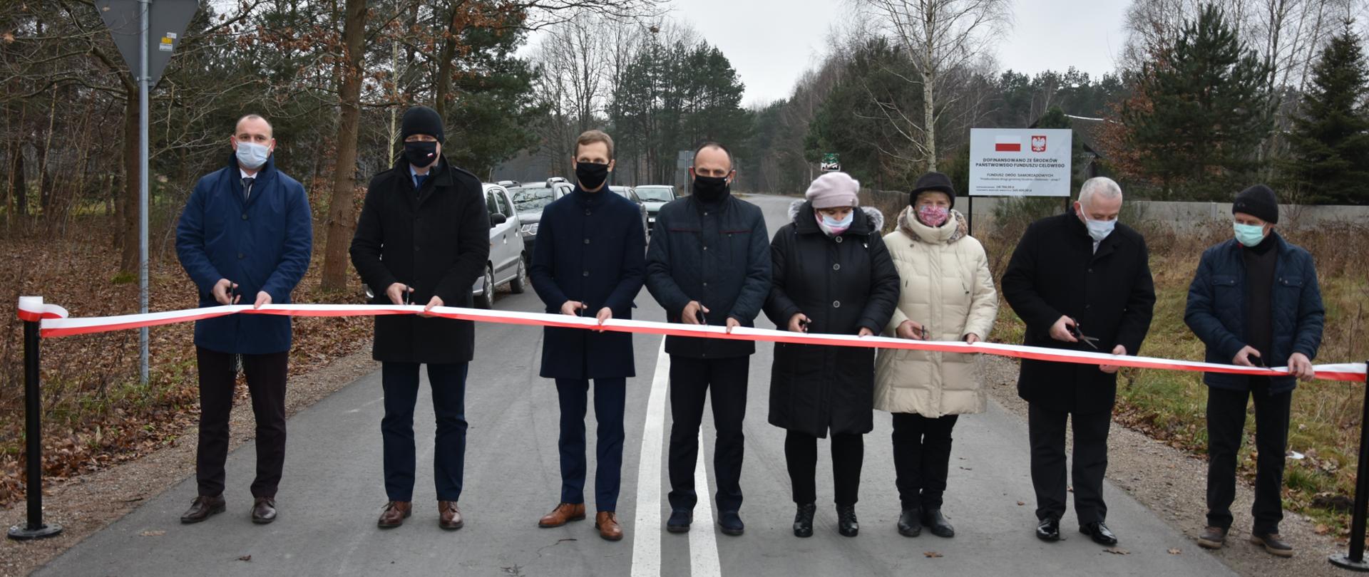 Zdjęcie przedstawia moment symbolicznego przecięcia wstęgi, po którym nastąpiło oficjalne oddanie do użytku fragment drogi gminna Studnie - Zieleniec. Wstęgę przecina sześciu mężczyzn i dwie kobiety. Osoby te stoją na zmodernizowanym fragmencie drogi, przed wstęgą - trzymając w rękach nożyczki. Fragment tej drogi przebiega przez obszar zalesiony.