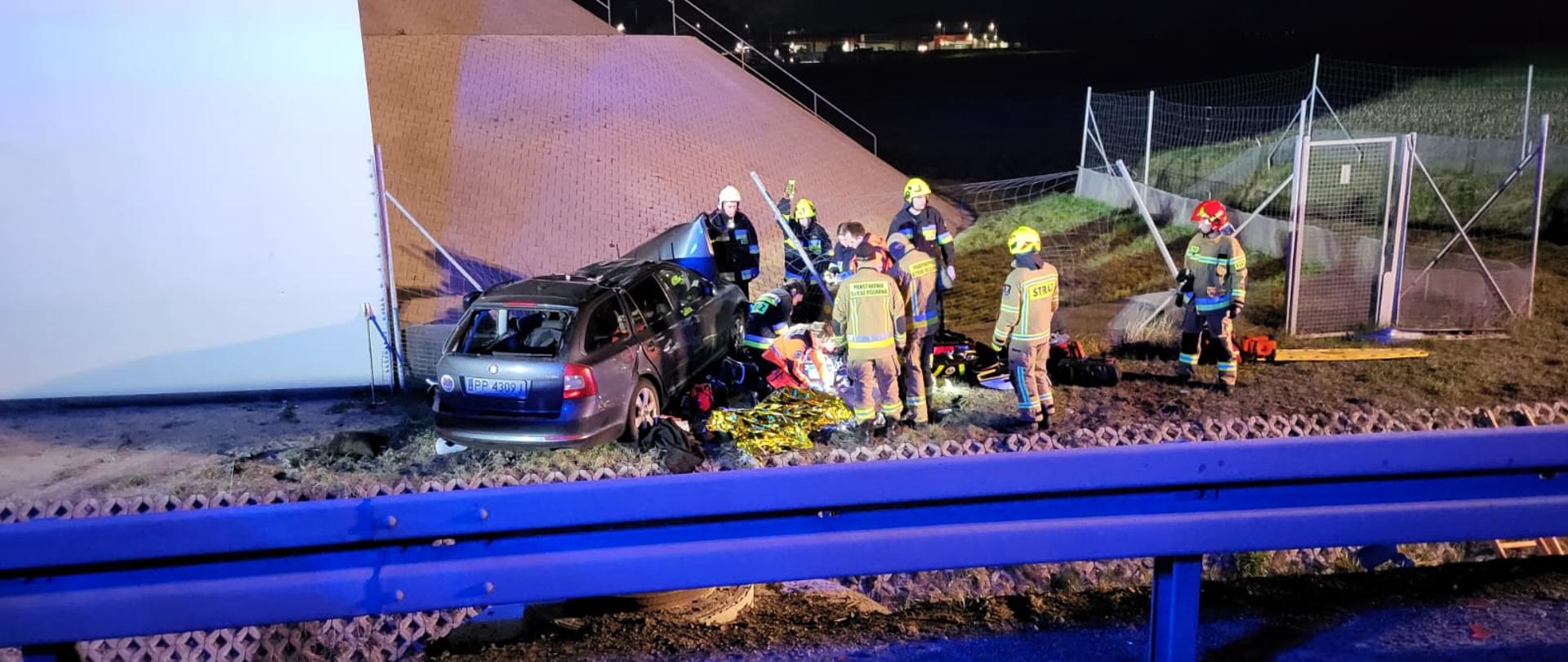 Uszkodzony samochód znajduje się na poboczu drogi obok pracujący strażacy. 