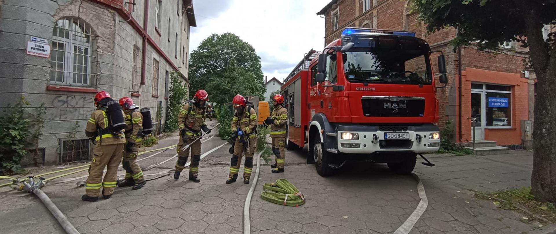 Samochód strażacki i strażacy przygotowujący linie gaśnicze oraz sprzęt przed wejściem do budynku