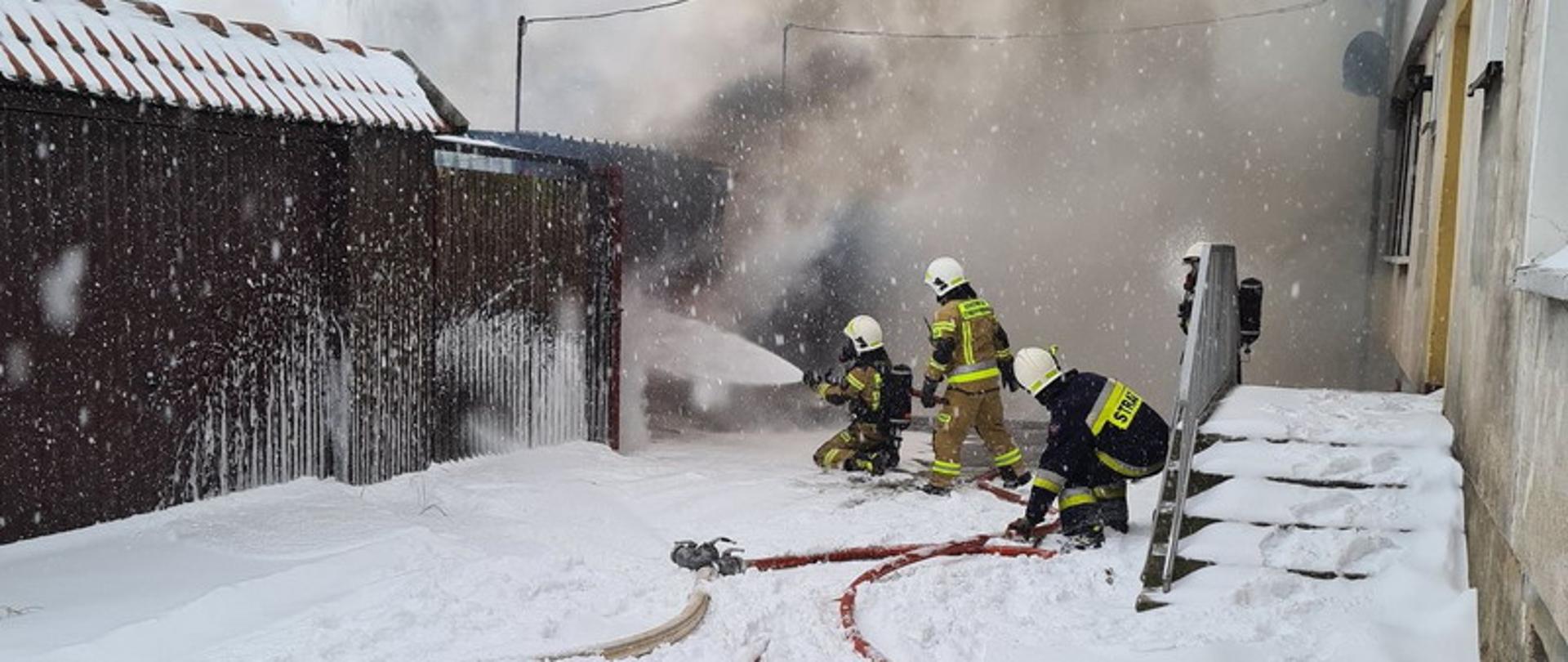 Na pierwszym planie dwa odcinki węży na dużej ilości śniegu. W tle widać heroiczną walkę strażaków podających prąd wody na palący się garaż, z którego wydobywają się kłęby ciemnego dymu. Po prawej stronie widać zaśnieżone wejście do bloku, a po prawej stronie szereg blaszanych garaży. 