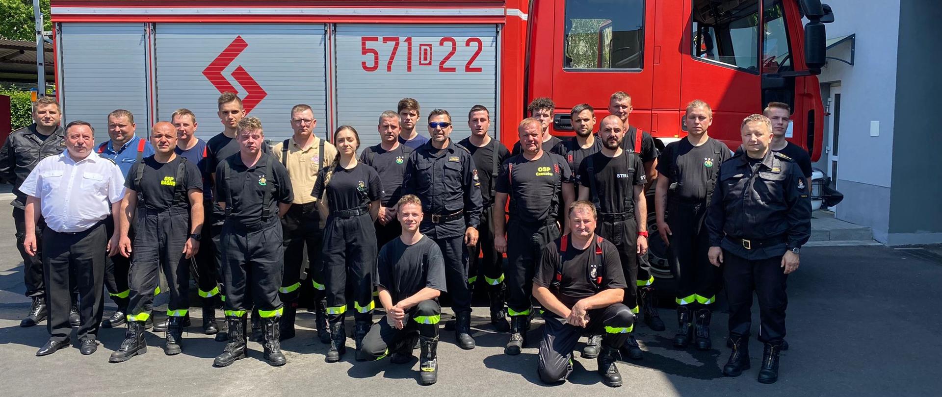 Zdjęcie grupowe. Na tle samochodu pożarniczego uczestnicy kursu wraz z funkcjonariuszami PSP. Wszyscy w czarnym umundurowaniu koszarowym.