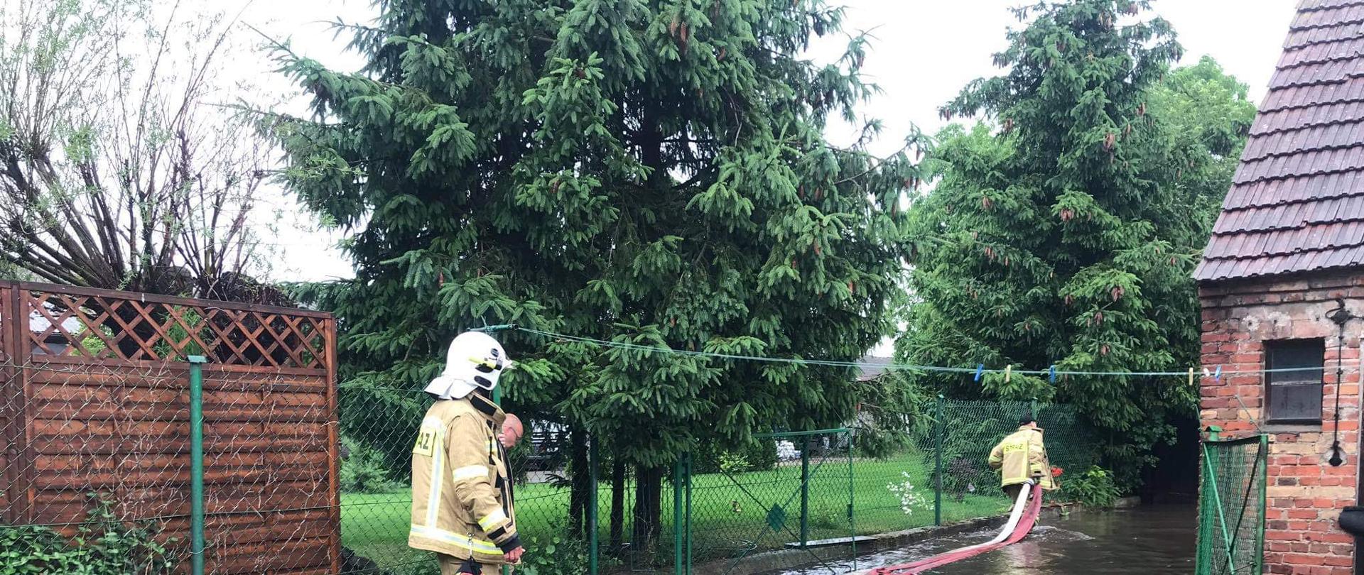Zdjęcie przedstawia zalana posesje oraz strażaków z pompą idacych w kierunkubudynku gospodarczego