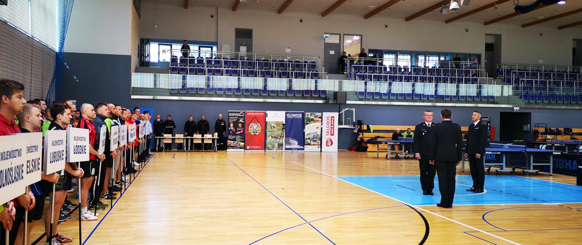 Zdjęcie przedstawia meldunek o gotowości zawodników do rozpoczęcia mistrzostw Zastępcy Śląskiego Komendanta Wojewódzkiego st. bryg. Mirosławowi Synowcowi składany przez komendanta zawodów mł. bryg. Jacka Szczypiorskiego. Strażacy podczas składania meldunku w ubraniach wyjściowych, pozostali strażacy w strojach sportowych. Zdjęcie wykonane na sali gimnastycznej.