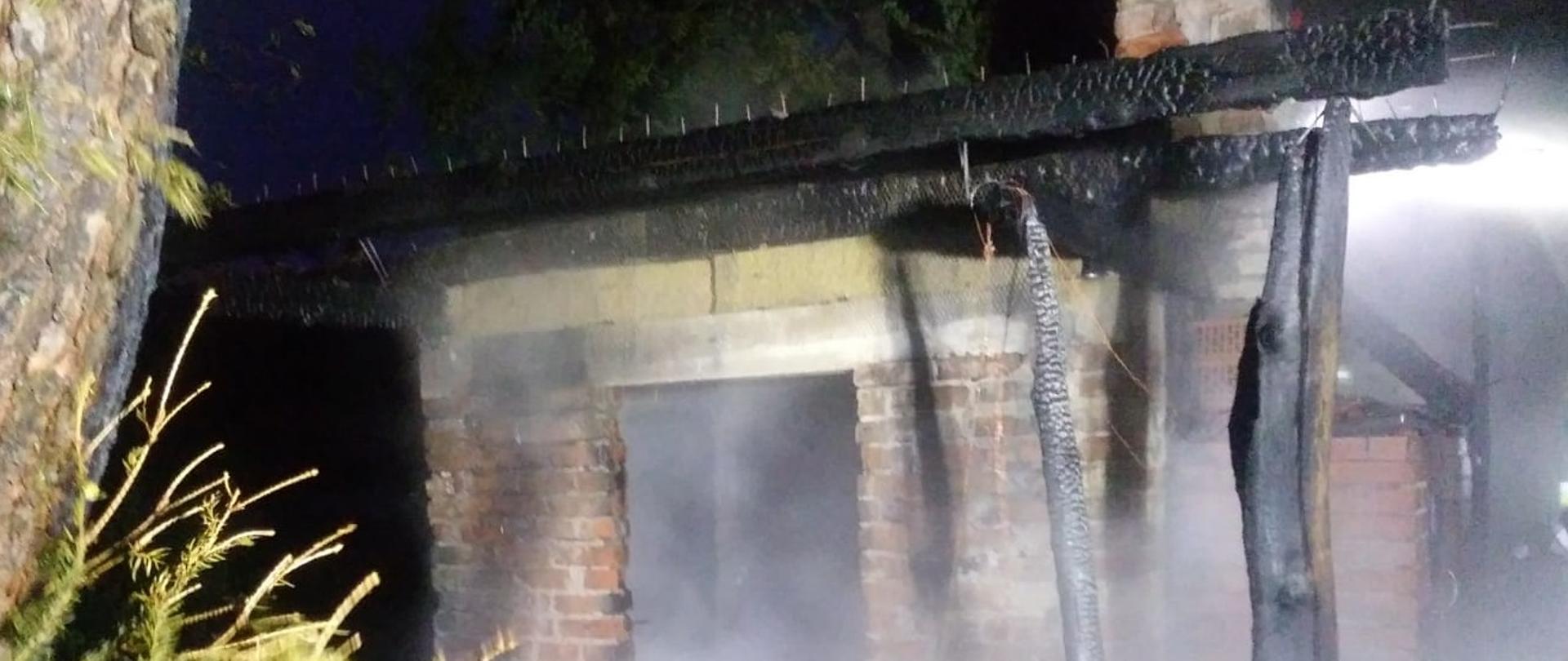 Pora nocna, spalony murowany budynek sauny ogrodowej. Nad nim widać spaloną konstrukcję dachu. 