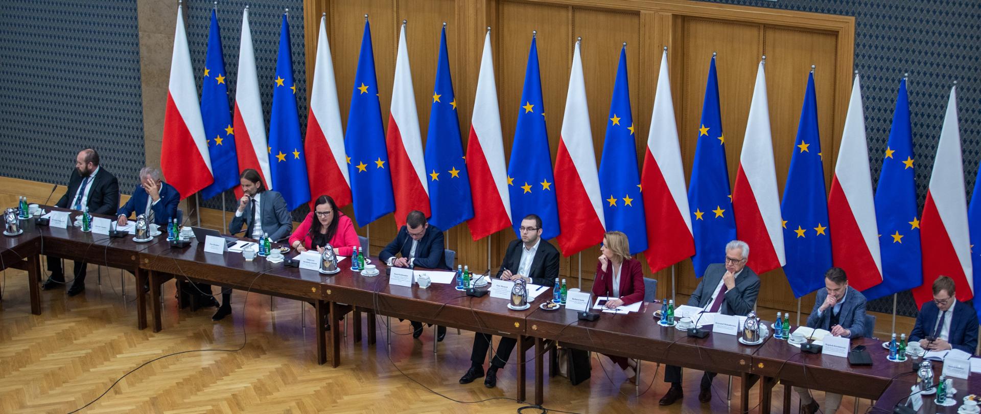 Na sali obrad przy długim stole siedzi grupa osób. Za nimi flagi Polski i Unii Europejskiej.