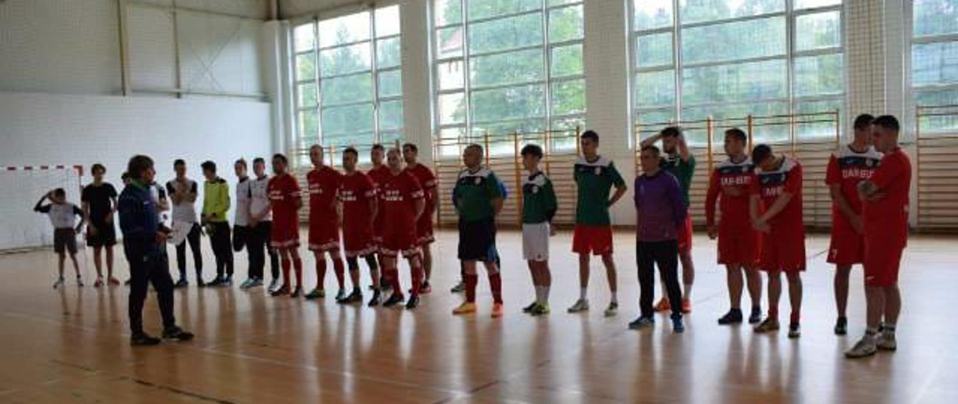 Zdjęcie przedstawia cztery drużyny podczas VIII Turniej Piłki Nożnej Służb Mundurowych o Puchar Wójta Gminy Rąbino stojące na hali sportowej.