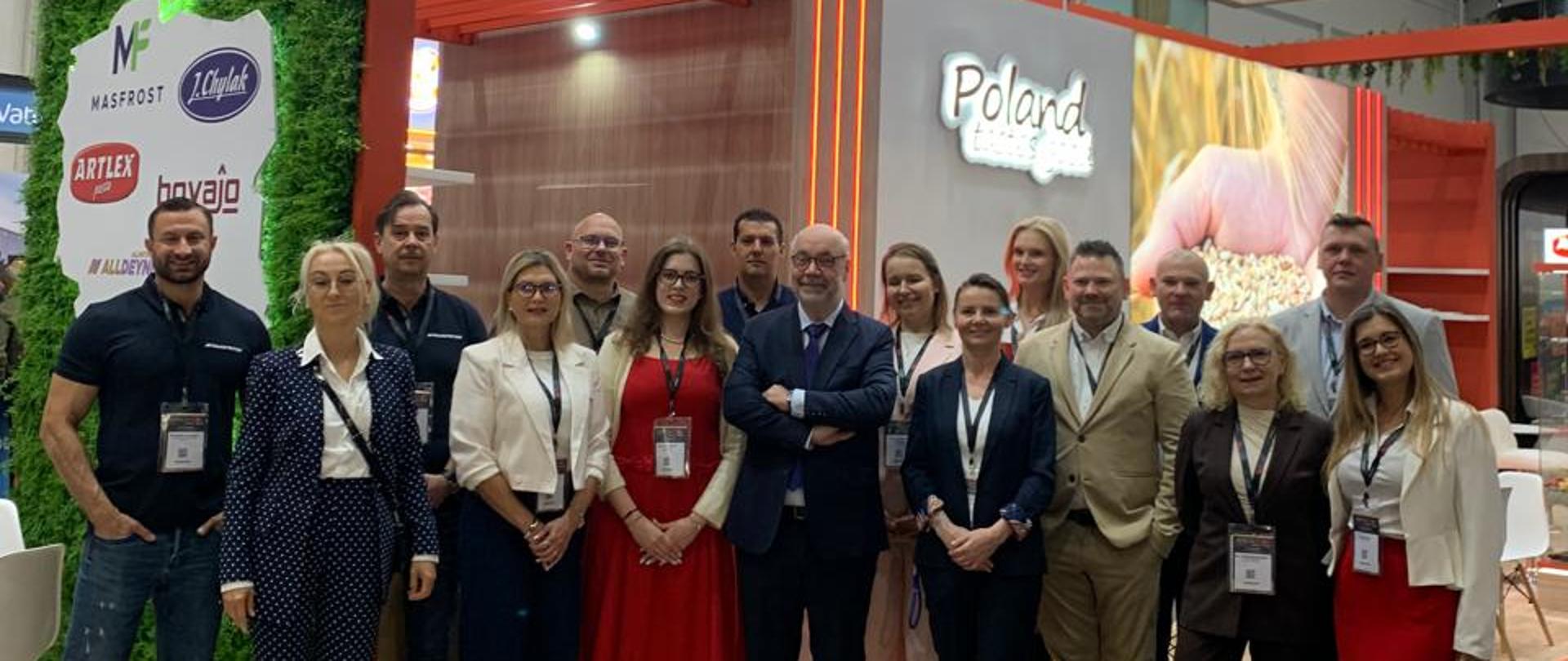 Zdjęcie grupowe 16 osób, wystawców polskich firm spożywczych; w centrum zdjęcia stoi Charge d'affaires Ambasady RP w Pretorii; w tle polskie stoiska i napis Poland oraz Poland Tastes Good; z lewej strony za grupą fotografowanych osób wisi mapa Polski z zaznaczonymi pięcioma firmami, wystawcami na targach Africa Food Show w Kapsztadzie. W wystroju wnętrza dominuje kolor pomarańczowy.