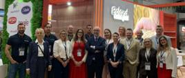Zdjęcie grupowe 16 osób, wystawców polskich firm spożywczych; w centrum zdjęcia stoi Charge d'affaires Ambasady RP w Pretorii; w tle polskie stoiska i napis Poland oraz Poland Tastes Good; z lewej strony za grupą fotografowanych osób wisi mapa Polski z zaznaczonymi pięcioma firmami, wystawcami na targach Africa Food Show w Kapsztadzie. W wystroju wnętrza dominuje kolor pomarańczowy.