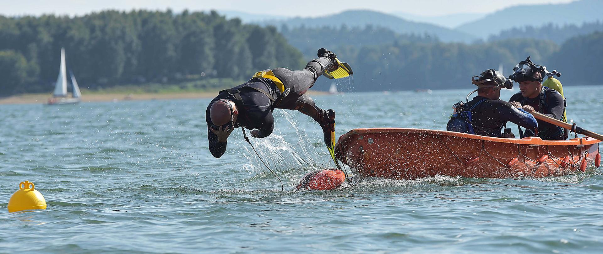 Zdjęcie zrobione na zewnątrz. Na pierwszym planie widać wodę i łódkę na której płynie trzech strażaków, założone mają na sobie ubrania nurkowe i sprzęt nurkowy. Jeden z nich wskakuje do wody. Na drugim planie w tle widać drzewa.