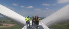 Na zdjęciu trójka strażaków ze Specjalistycznej Grupy Ratownictwa Wysokościowego z Radzionkowa ubrani w specjalistyczny kombinezon przeznaczony do działań na wysokości oraz uprzęże, które obejmują swoim zakresem biodra oraz ramiona strażaka wyposażeni w lekkie kaski przeznaczone do prac na wysokości wraz z przedstawicielem Farmy Wiatrowej prowadzącym szkolenie ubranym w specjalistyczny kombinezon oraz kask przeznaczony do prac na wysokości . Na szczycie turbiny wiatrowej. W tle łopaty turbiny oraz widok z góry na horyzont gdzie widać pola, domy, las i niebo.